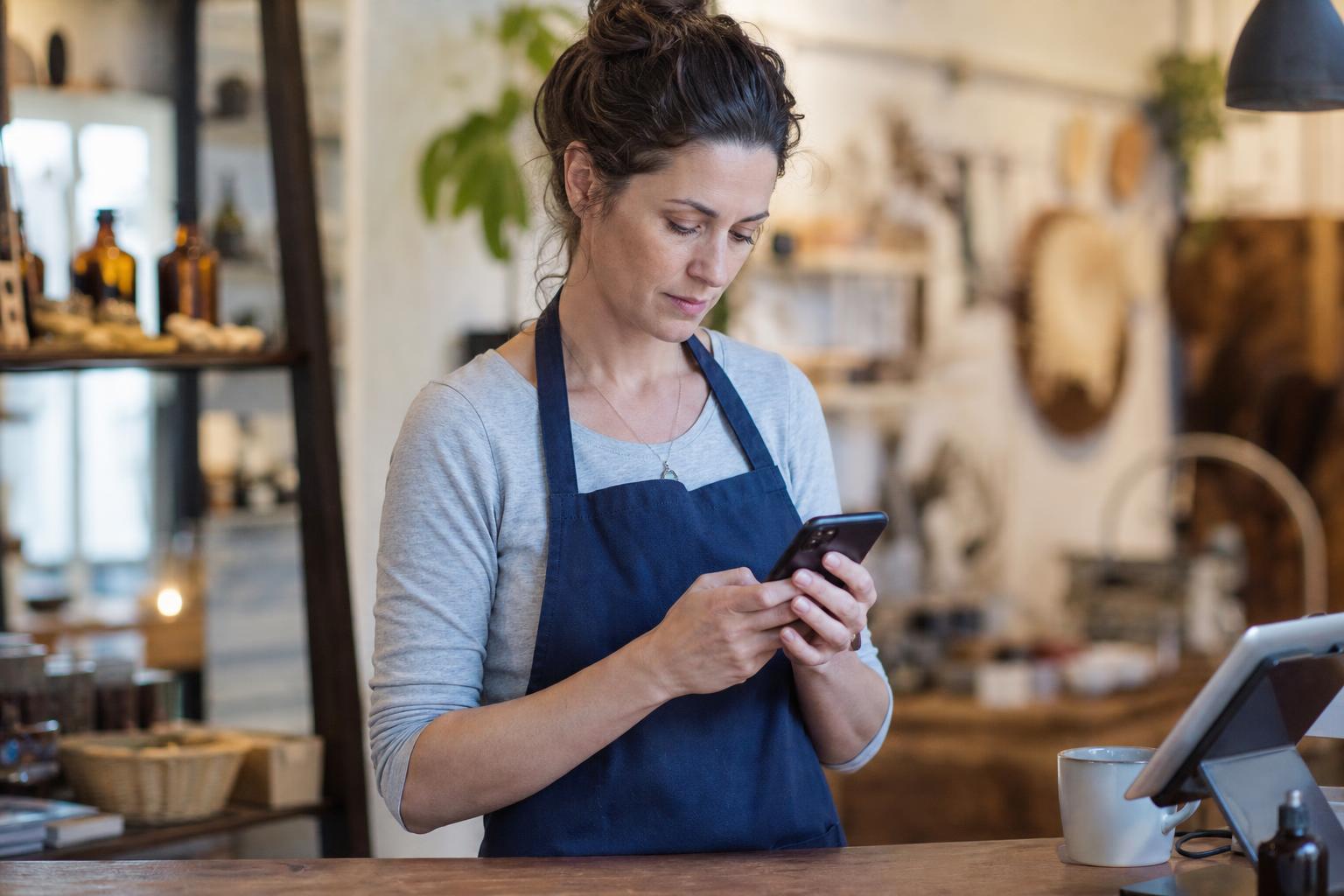 A small business owner standing behind a shop counter typing a reply to a customer review on a smartphone