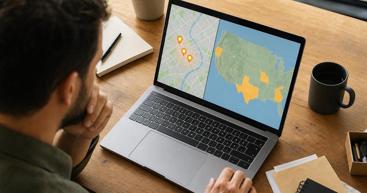 A small business owner at a wooden desk looking at a laptop screen split between a close-up local city map with pin markers and a wider country-level map with highlighted regions